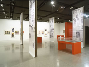 View of exhibition showing panels of newspaper collages, documents in display cases and poster art.